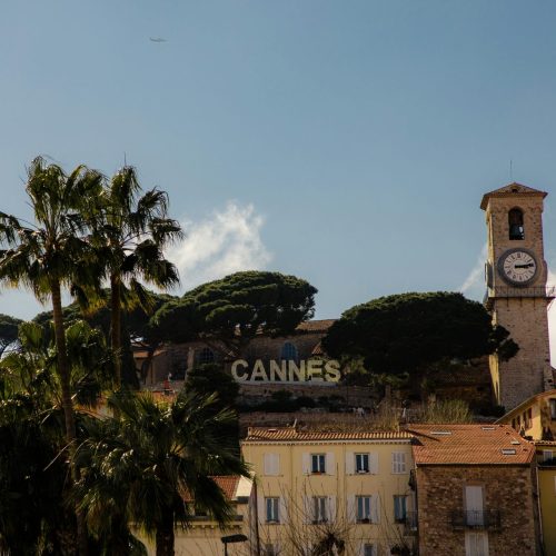 Vue sur « Cannes » au sommet de la ville avec une église en pierre, des palmiers et des pins avec des maisons colorées jaune/orange en contre bas