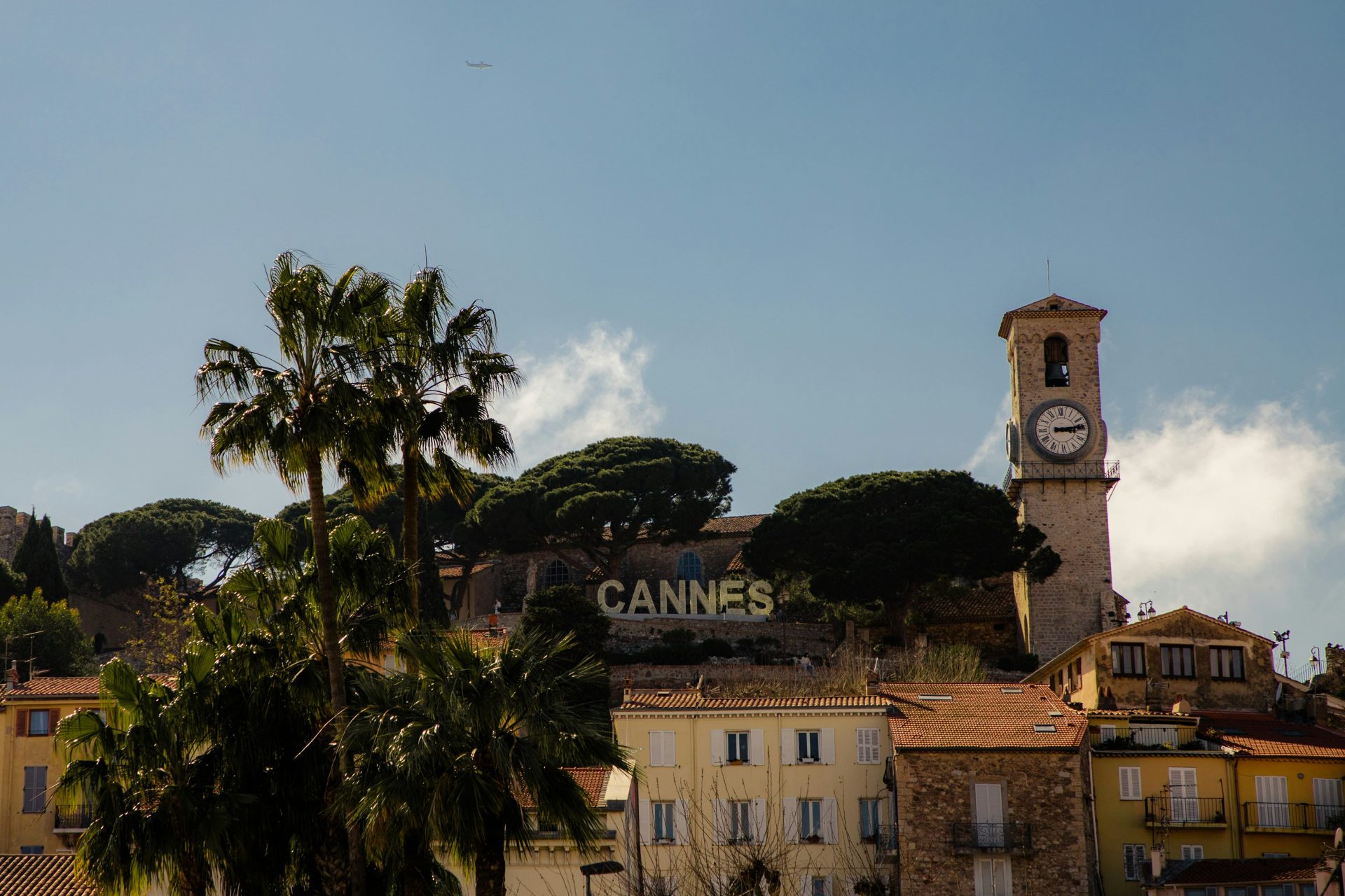 Vue sur « Cannes » au sommet de la ville avec une église en pierre, des palmiers et des pins avec des maisons colorées jaune/orange en contre bas