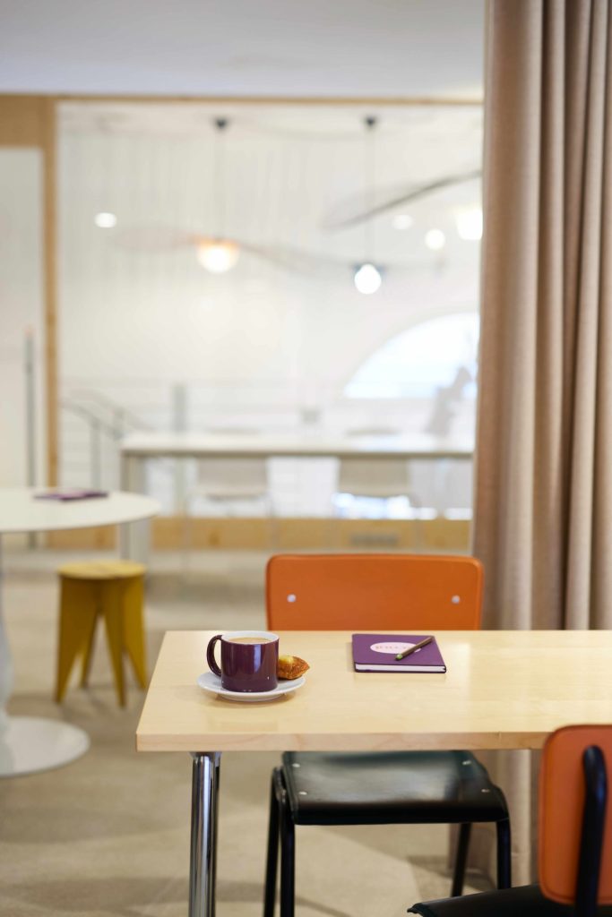 Bureau en bois clair avec une tasse à café et une viennoiserie, un carnet violet scool ainsi qu’un stylo posé sur la table 