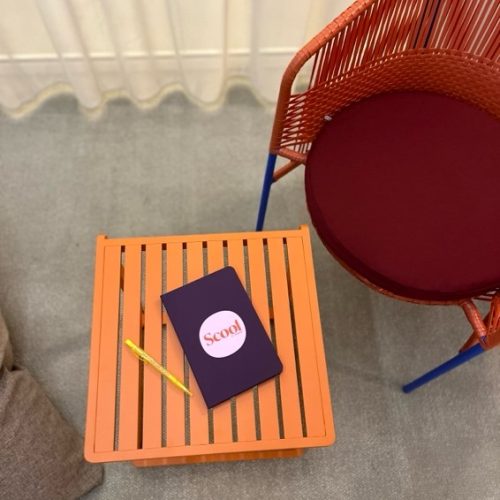 Vue de haut d’une table basse orange avec un carnet violet et rose avec pour inscription « Soniho » dessus et un crayon à papier jaune.