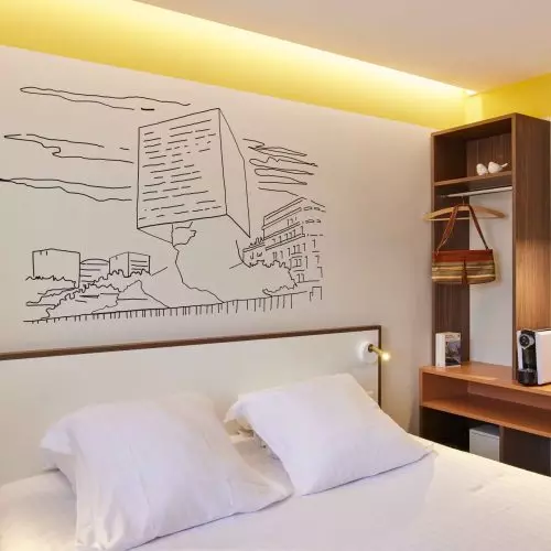 Chambre de l’hôtel du pin à Nice avec un pan de mur avec une illustration filaire noir de la tête carrée à Nice, des équipements de la chambre, une tête de lit, une étagère en bois, une machine à café, des tasses à café, un luminaire pendant et un lit double